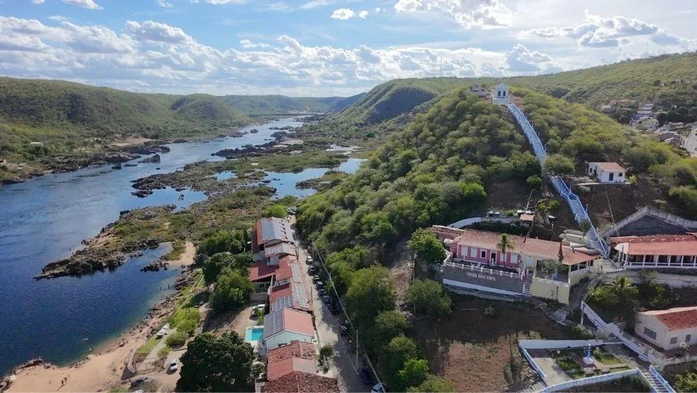 Vista aérea da cidade de Piranhas, mostrando o Rio São Francisco serpenteando entre formações rochosas e áreas de vegetação típica do sertão. À direita, um morro com casas coloridas e uma longa escadaria branca que leva até o topo, onde há uma pequena construção. Na parte inferior, próximas ao rio, aparecem edificações com telhados vermelhos, algumas árvores e uma faixa de areia na margem. O céu está azul, com nuvens espalhadas, iluminando a paisagem.